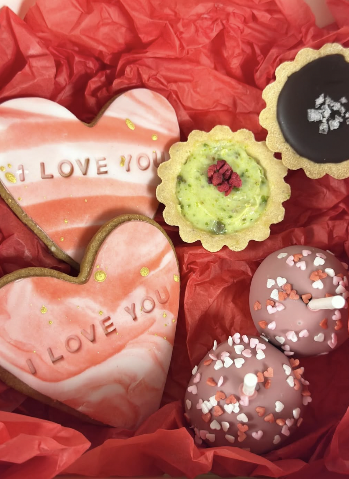 Valentine's Day cookies and cake pops on red tissue paper. Two heart-shaped cookies with pink and white marbled icing and the words 'I Love You' written on them. Two small tartlets, one with green filling and pink topping, and the other with dark cho