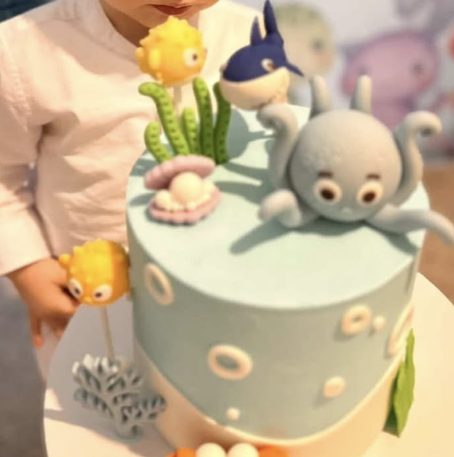 A children's birthday cake decorated with colorful ocean-themed fondant figures, including a gray octopus with big eyes, a blue whale, and orange and yellow fish. A child is holding a fish-shaped cake topper.