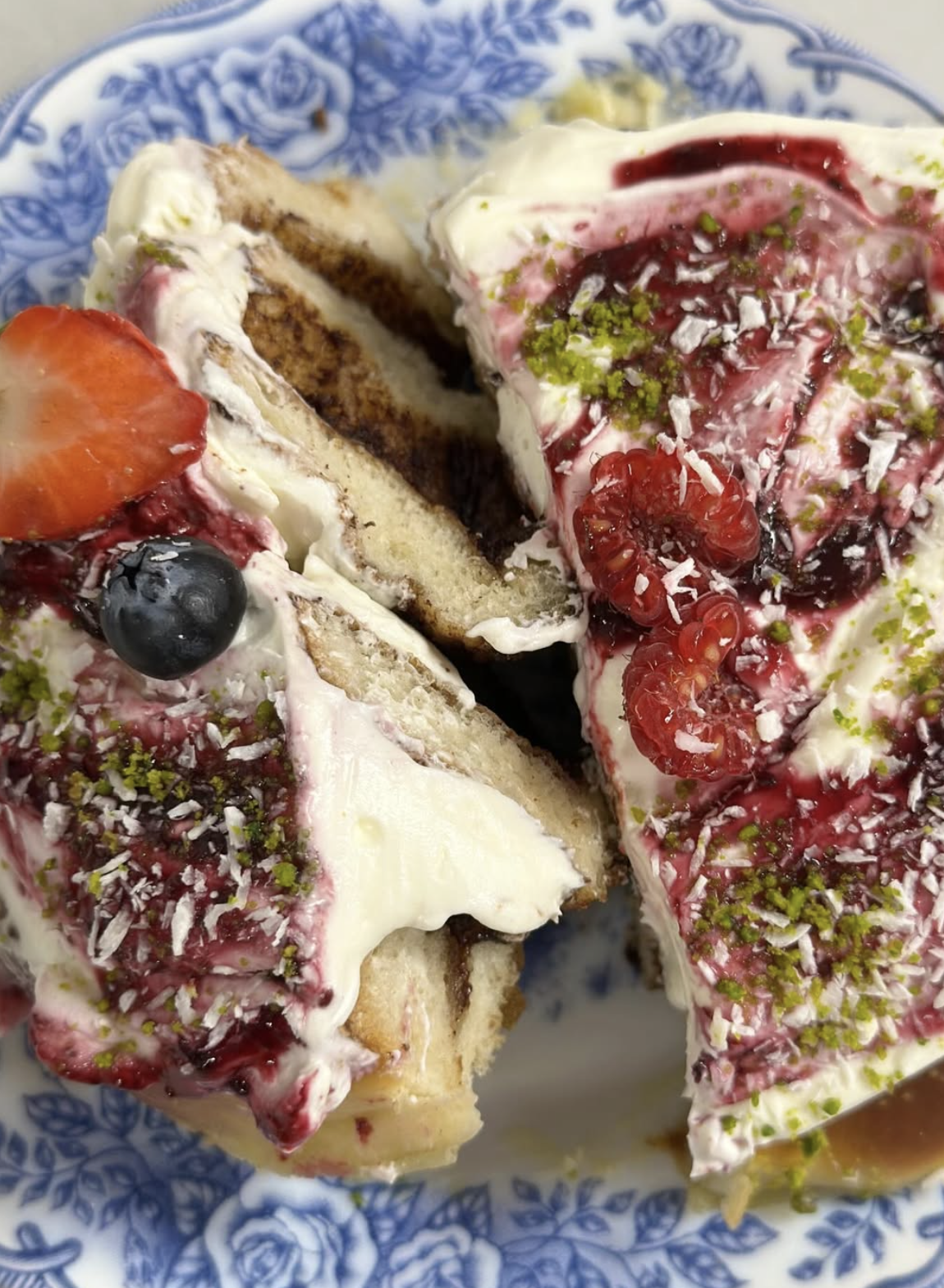 A close-up of a cake slice with layers of white cake, cream, and berry filling topped with fresh berries, shredded coconut, and chopped pistachios on a blue and white patterned plate.