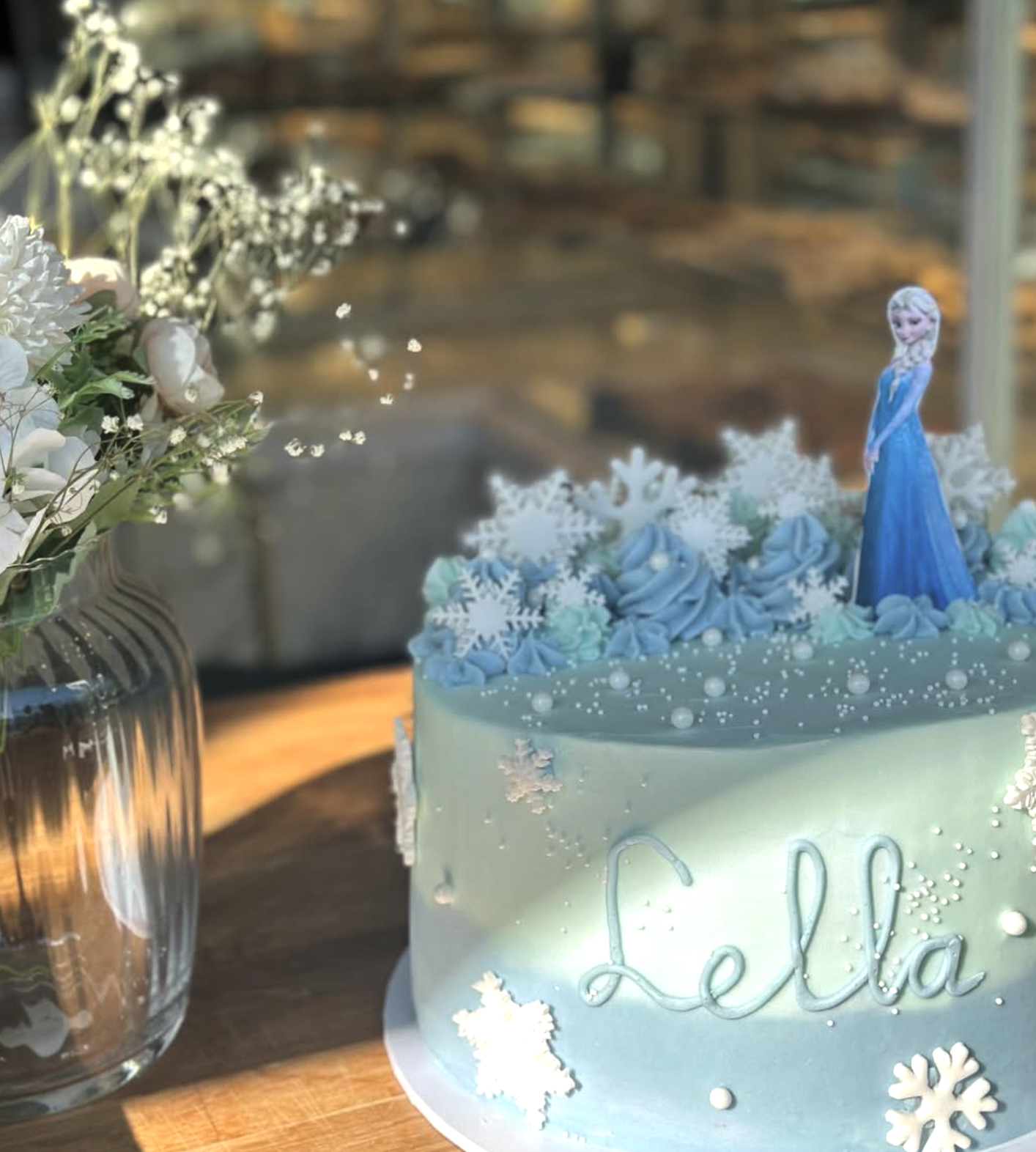 A birthday cake decorated with snowflakes and blue flowers, featuring a figurine of Elsa from Frozen, with the name 'Lella' written on the front, placed on a wooden table beside a glass vase with white flowers.