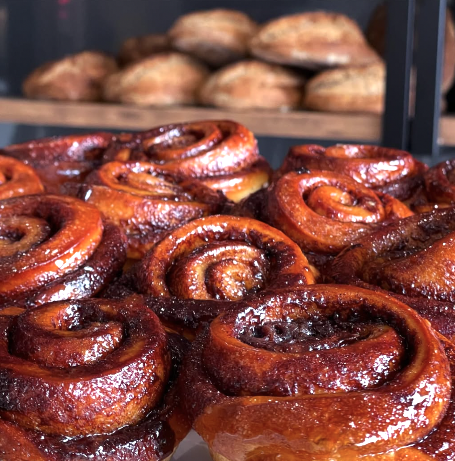 Close-up of glazed cinnamon rolls in a bakery display case, with some baked goods in the background.