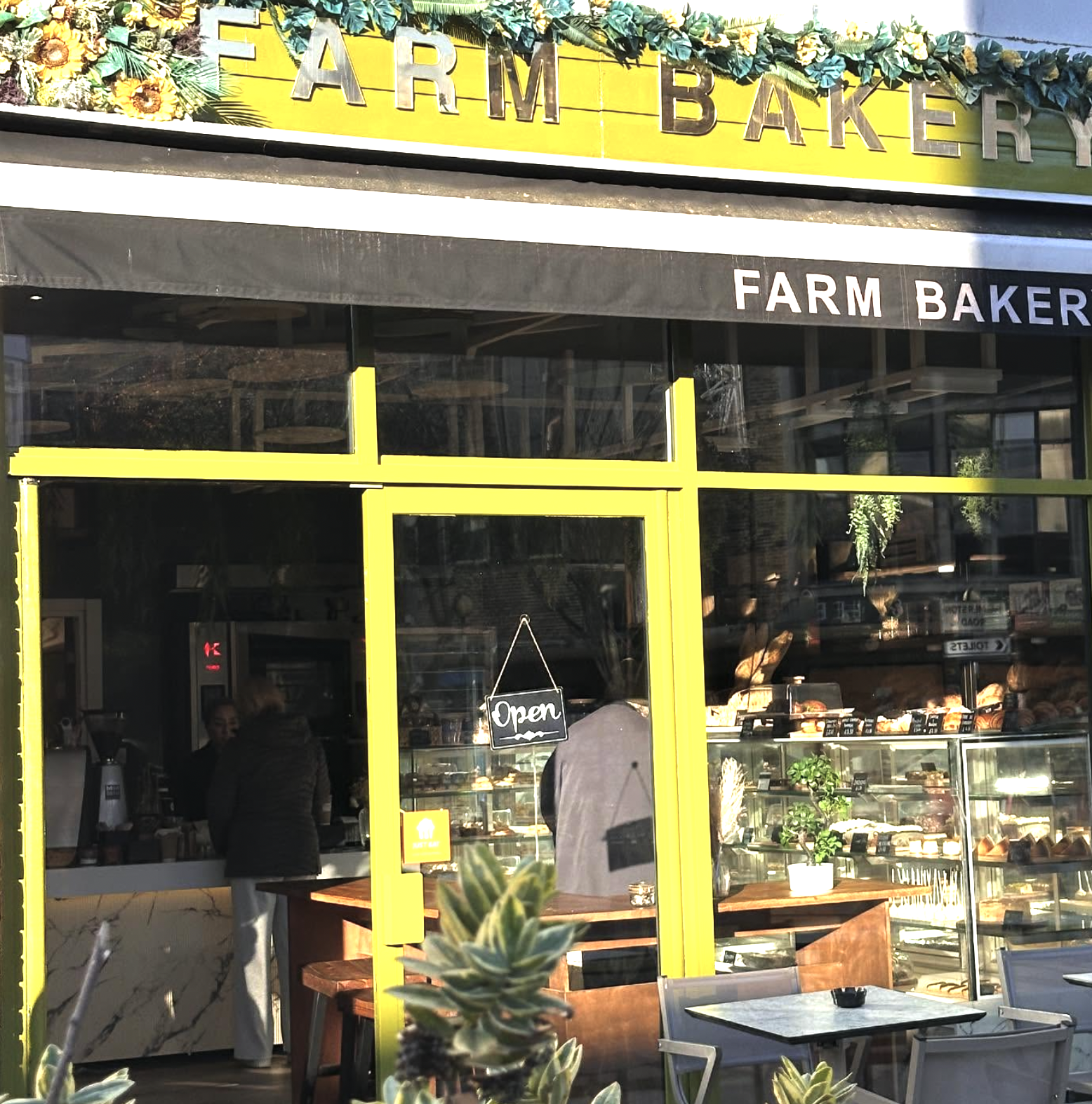 The storefront of a bakery named 'Farm Bakery,' with a yellow frame entrance, a black canopy, and a glass window showing baked goods inside. There is a small 'Open' sign hanging on the door, and some plants outside.