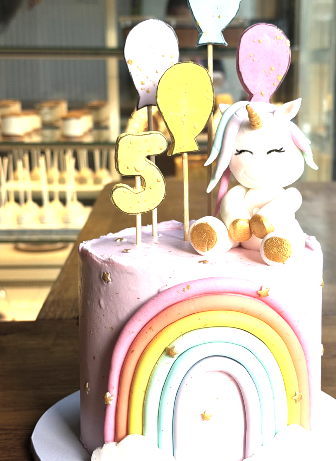 A pink birthday cake decorated with a rainbow and small star-shaped decorations, topped with a white unicorn figurine, colorful balloons, and a yellow '5' candle.