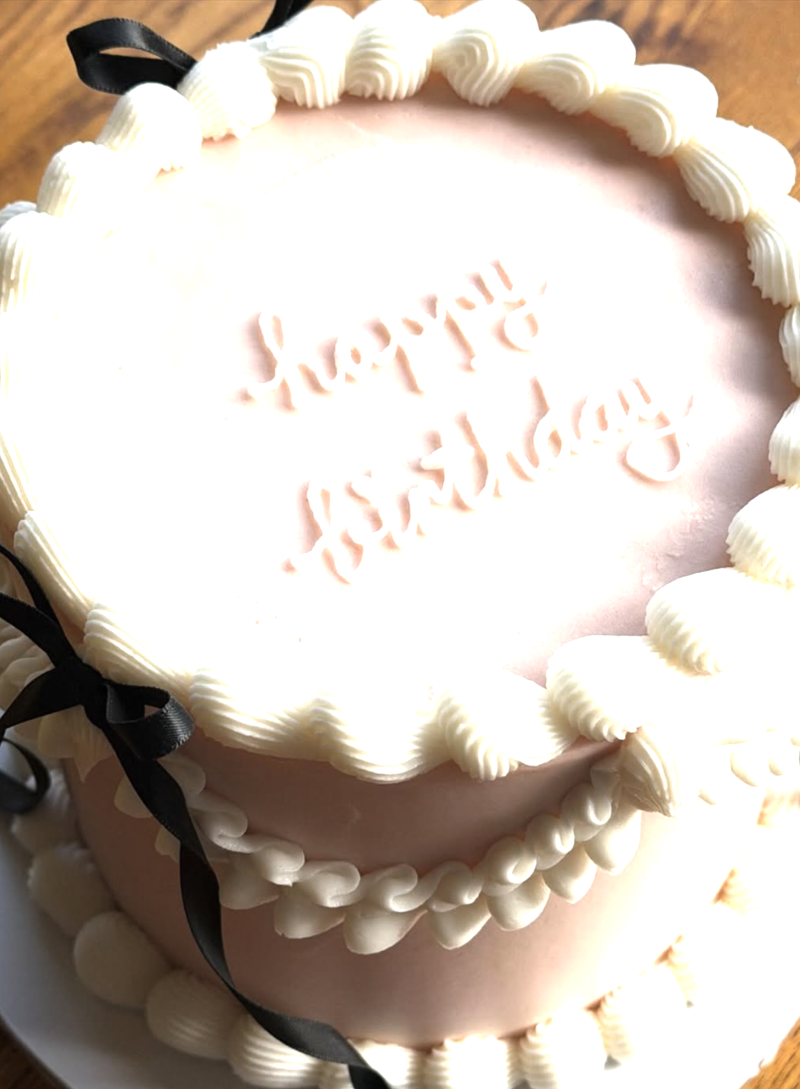 A round birthday cake with pink and white icing, decorated with white piped swirls around the edges and black ribbons on the side. The cake has a message written in pink icing that says "Happy Birthday."