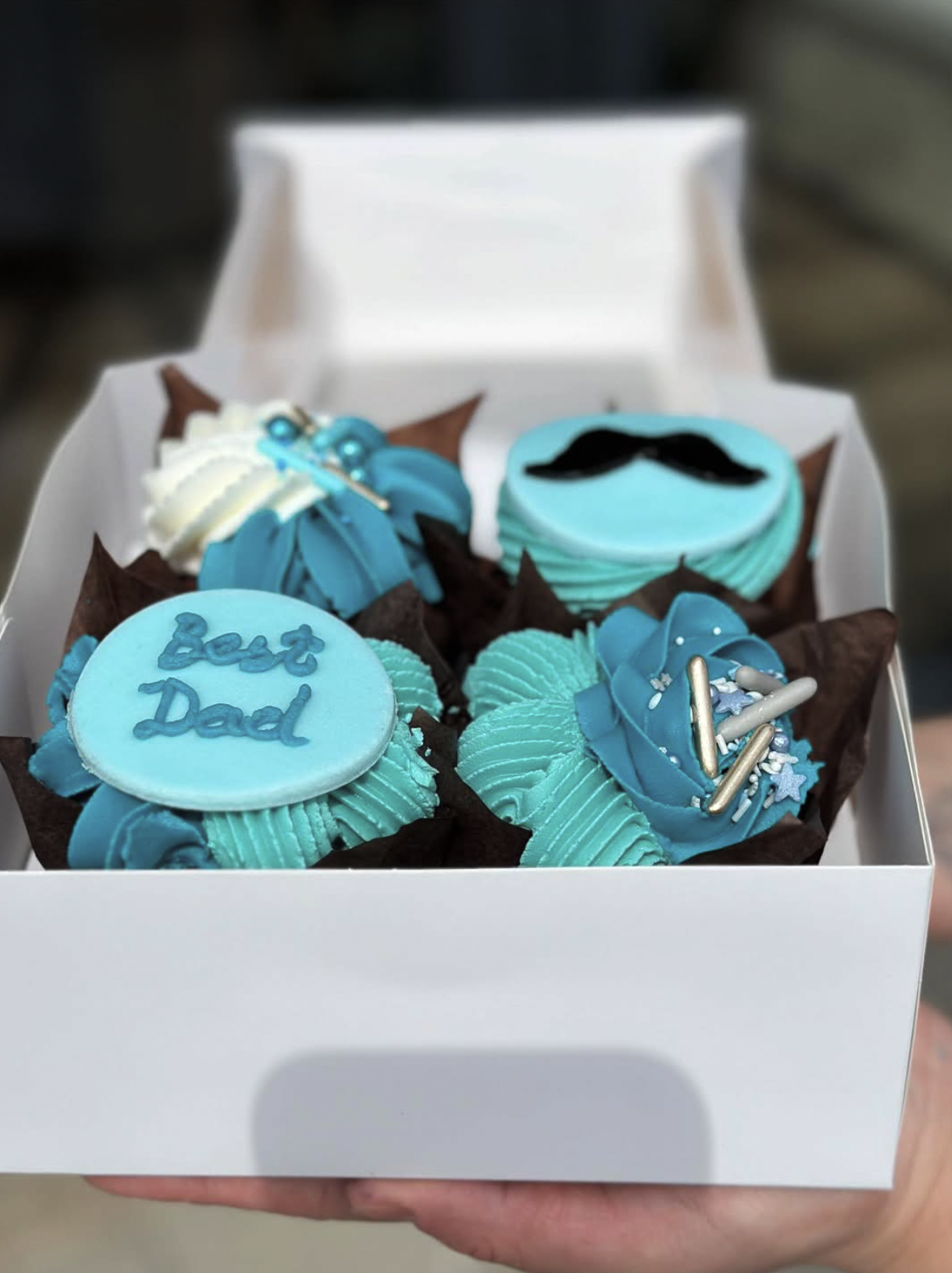 A white box of cupcakes decorated in blue and white, with one cupcake featuring the words "Best Dad," a mustache, and other decorations for a celebration.