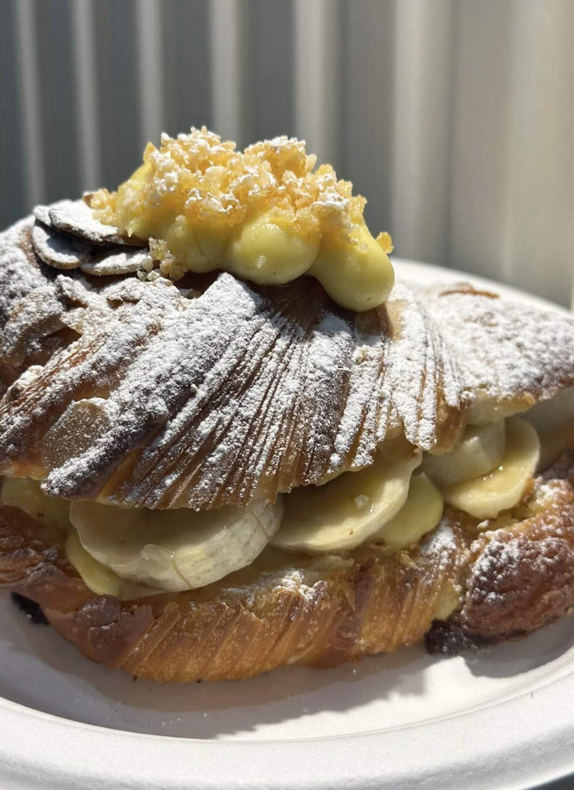 Close-up of a pastry topped with powdered sugar, banana slices, and creamy filling, served on a white plate.