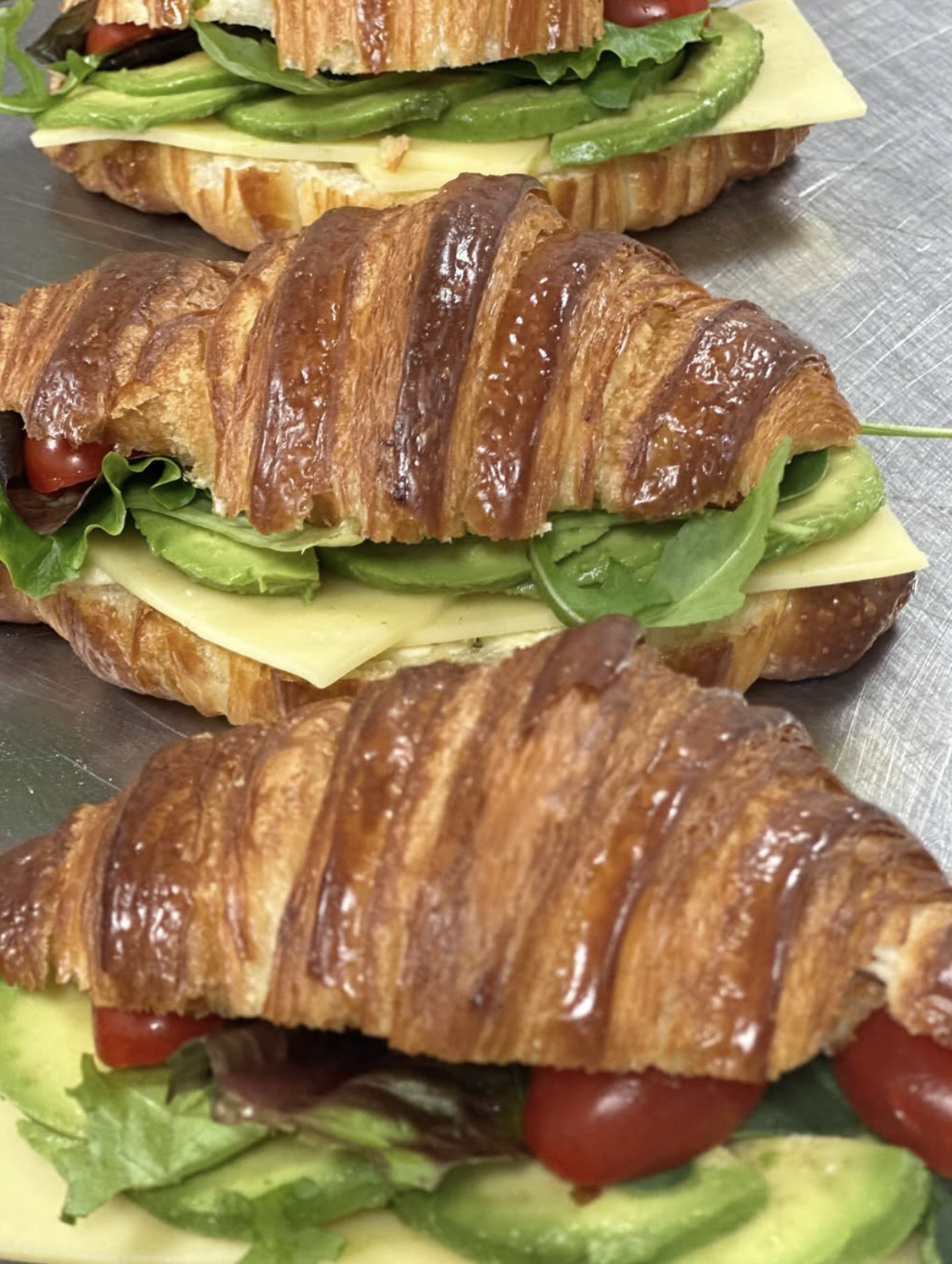 Three croissant sandwiches filled with lettuce, cheese, tomatoes, and sliced green bell peppers on a metal tray.