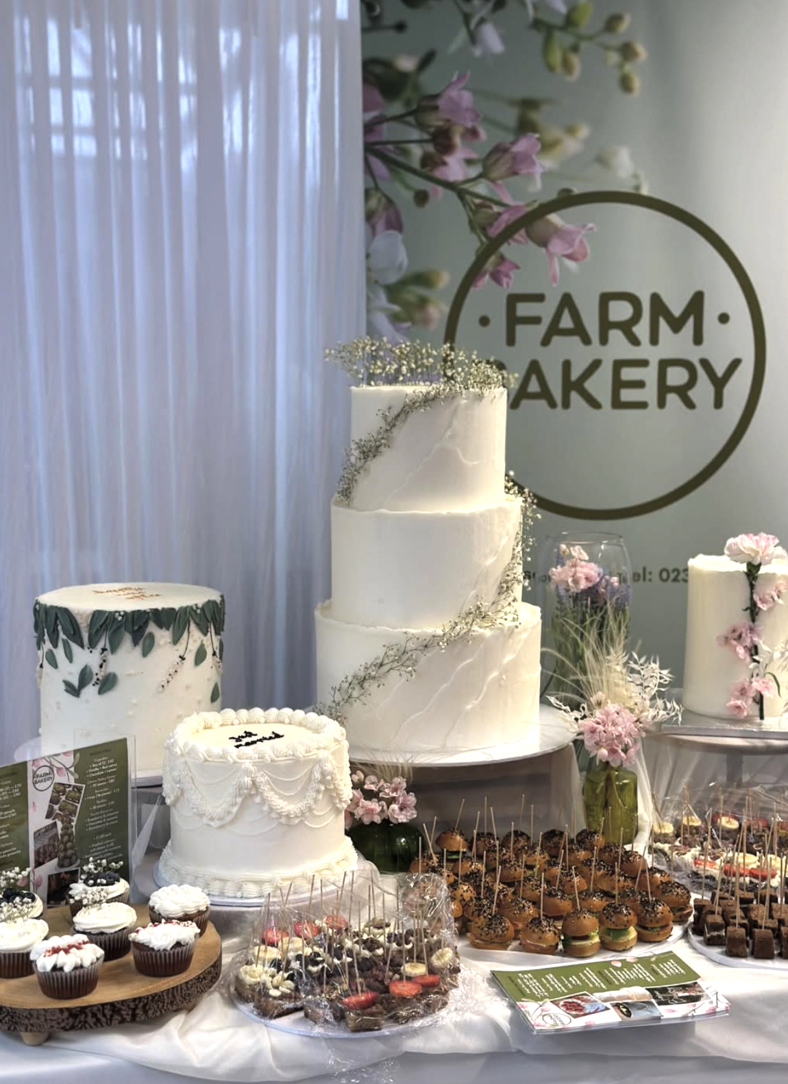 Display of various cakes and desserts on a table at an event, with a backdrop showing the 'Farm Bakery' logo and pink flowers for decoration.