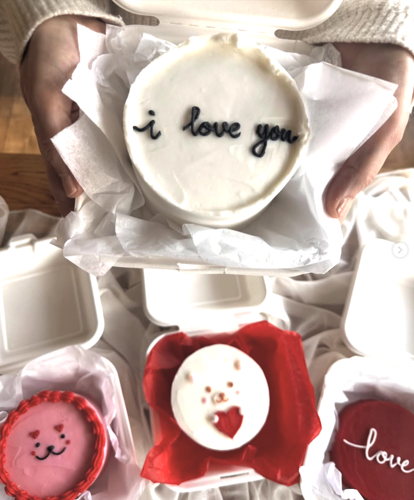 A person holding a small white cake with the words 'i love you' written on it in black icing. The cake is in a white box with tissue paper. There are other decorated cakes around, including a pink one with a cat face, a white one with bear features, 