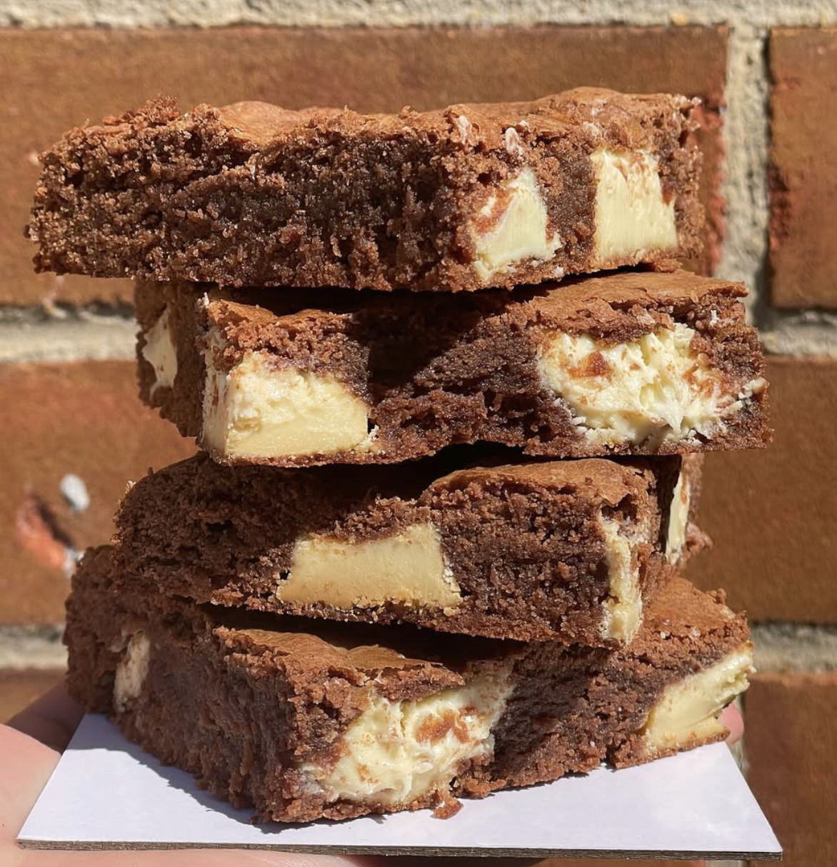 Stack of four brownie bars with white chocolate chunks, placed on white paper against a brick wall background.