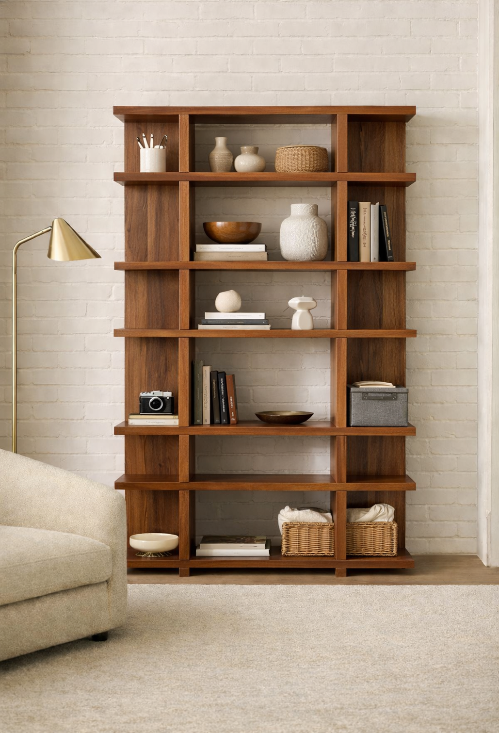A tall wooden bookshelf against a white brick wall with decorative vases, books, a vintage camera, baskets, and small sculptures, partially next to a beige sofa and a gold floor lamp in a cozy living room.