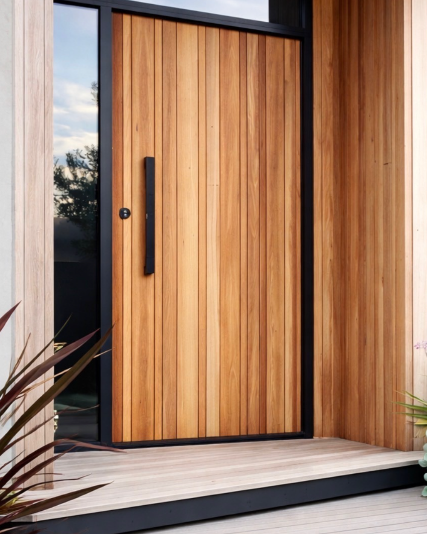 Modern front door made of wooden planks with black handle and black frame, surrounded by wooden siding and a small deck with plant in the foreground.