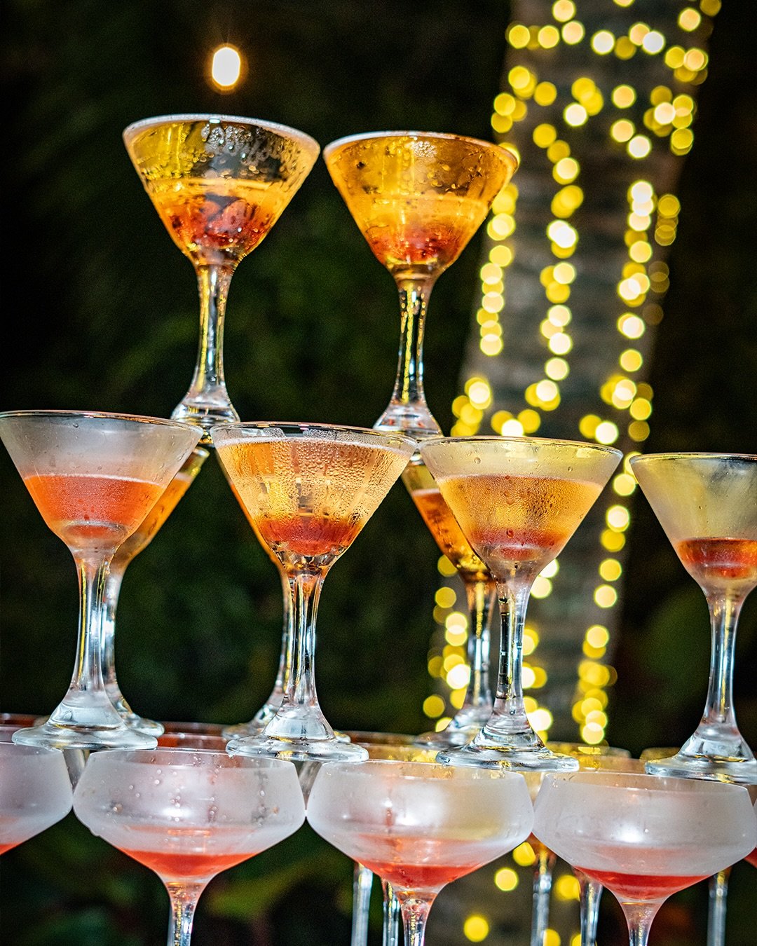 Stacked glasses with champagne or sparkling wine on a table at night with blurred yellow string lights in the background.