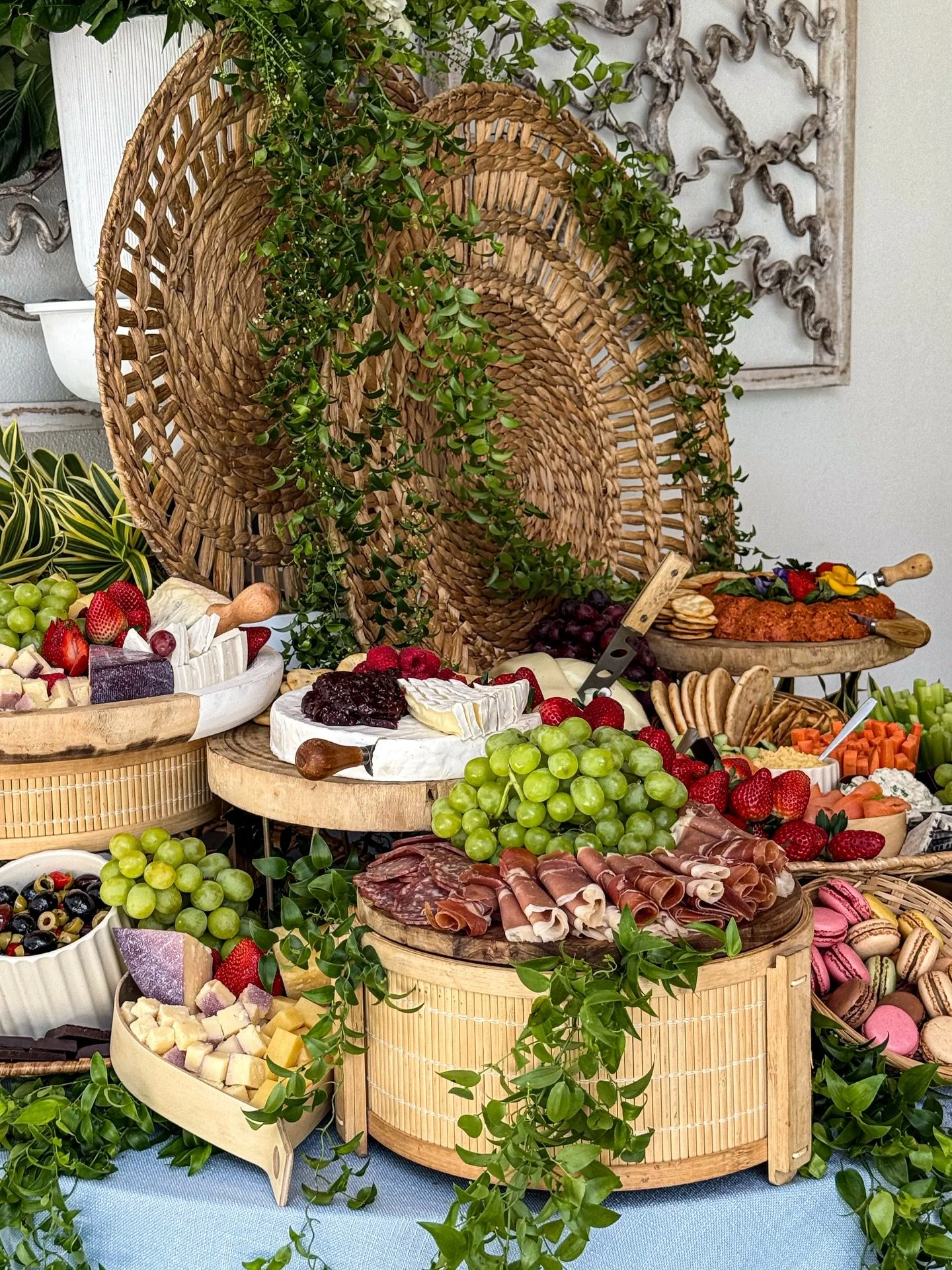 A cheese and charcuterie spread with grapes, strawberries, macarons, and other snacks on a table decorated with greenery.