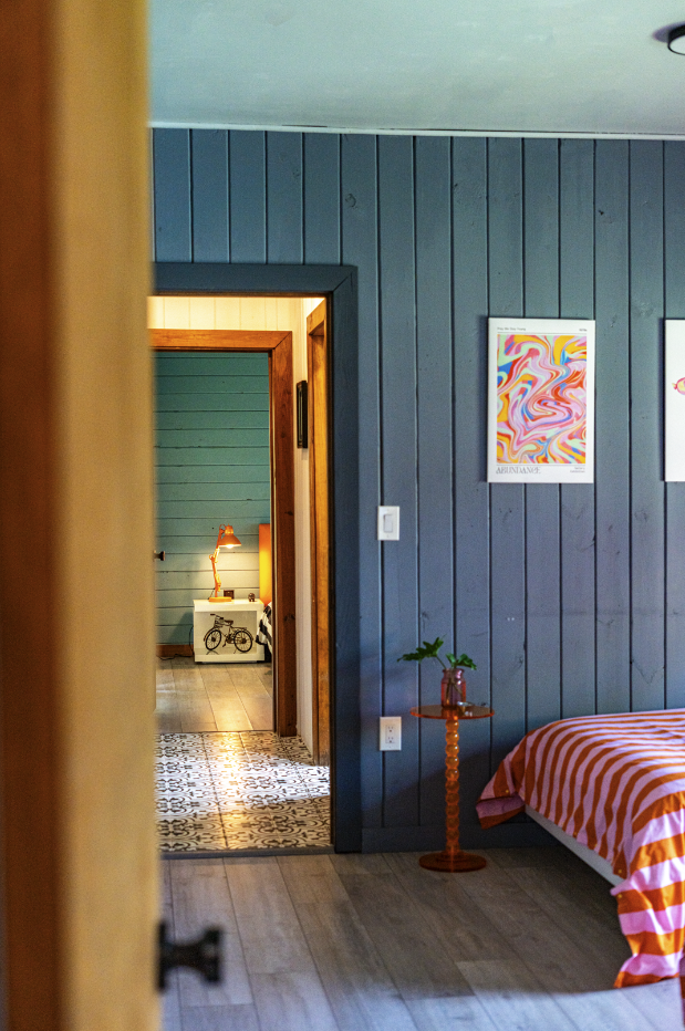Part of a bedroom with a bed and striped bedding, a small pink table with a plant, and artwork on the blue wall. A doorway leads to another room with a nightstand, lamp, and patterned tiles.
