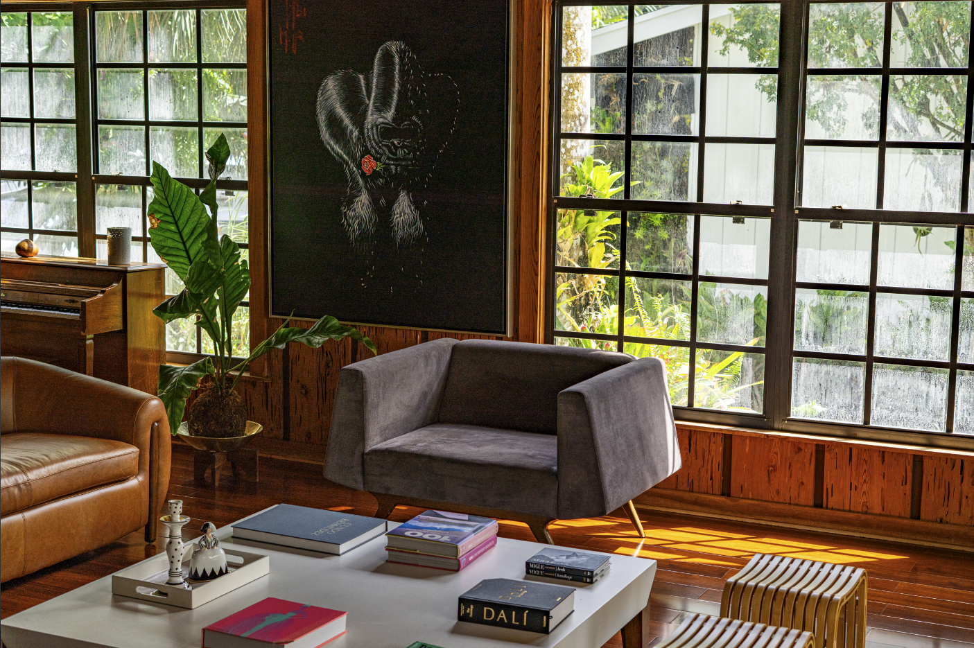 Living room with large windows, gray armchair, brown leather sofa, potted plant, white coffee table with books, a small wooden bench, and a framed black chalkboard with a drawing of a dog on the wall.