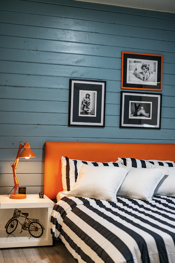 A bedroom with a blue wooden wall, three black-and-white framed photographs, an orange headboard, a white nightstand with a black bicycle decoration, an orange desk lamp, and a bed with black and white striped bedding and pillows.
