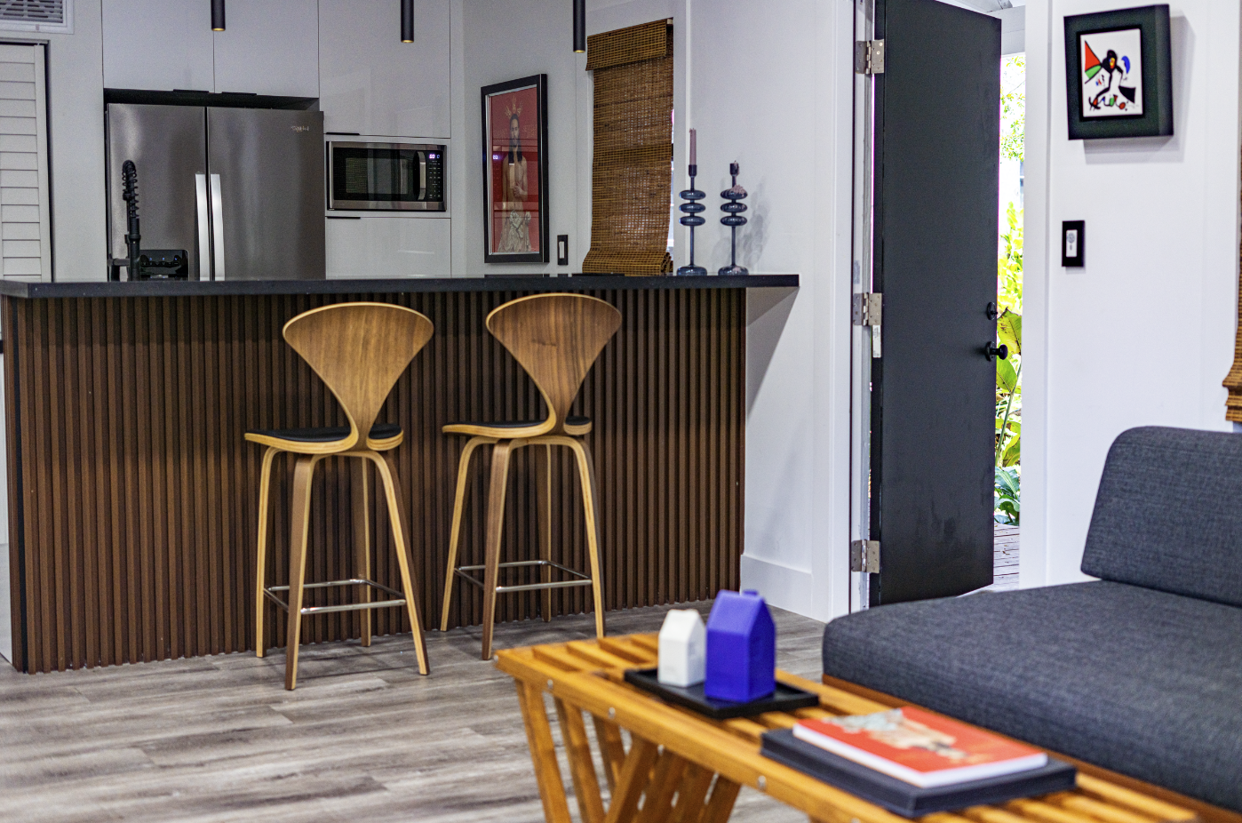 A modern living room with a kitchen area visible in the background. The kitchen features a stainless steel refrigerator, microwave, and white cabinetry. Two wooden bar stools are at the kitchen counter, and there are decorative candles on top. To the