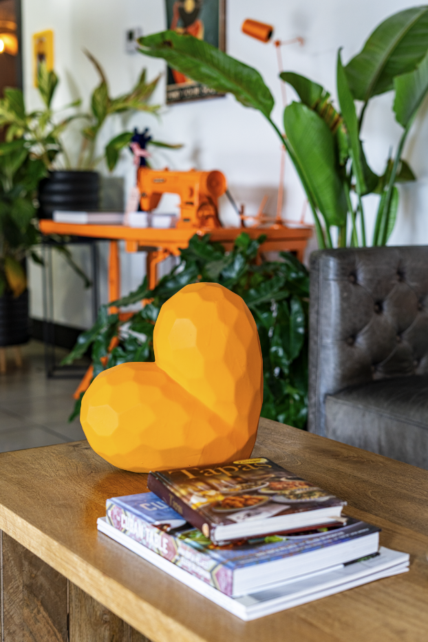 A yellow, faceted heart-shaped sculpture on a wooden table surrounded by magazines, with houseplants and an orange sewing machine in the background.