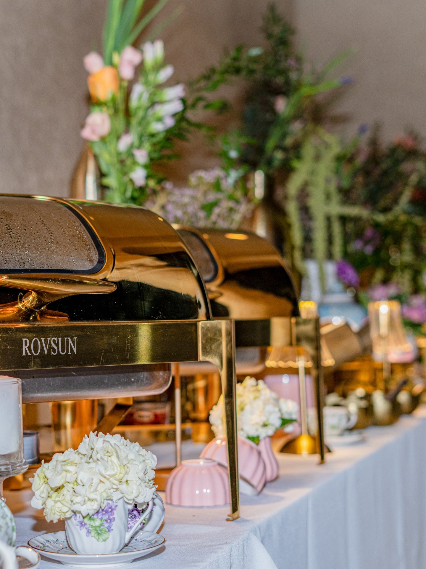 Chafing dishes on a buffet table decorated with flowers and candles, set against a backdrop of floral arrangements.