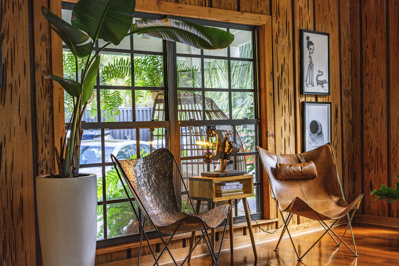 Interior of a cozy room with wooden walls, a large window, tropical plants, two leather chairs, a small wooden side table, and framed black and white art.