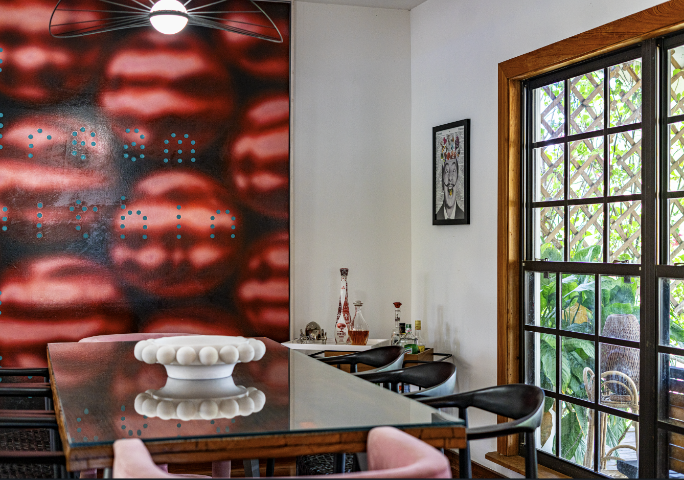 Interior of a dining area with a glass-top wooden table, black chairs, and a white decorative bowl, featuring a large abstract art piece of red lips on the wall, a framed black and white portrait, and a window with a view of greenery outside.