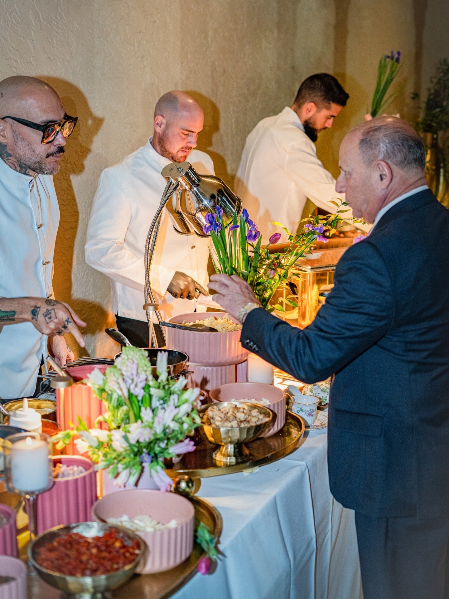A man in a black suit choosing food from a buffet table decorated with pink and gold accents and flowers, with three chefs in white jackets preparing food behind the table.