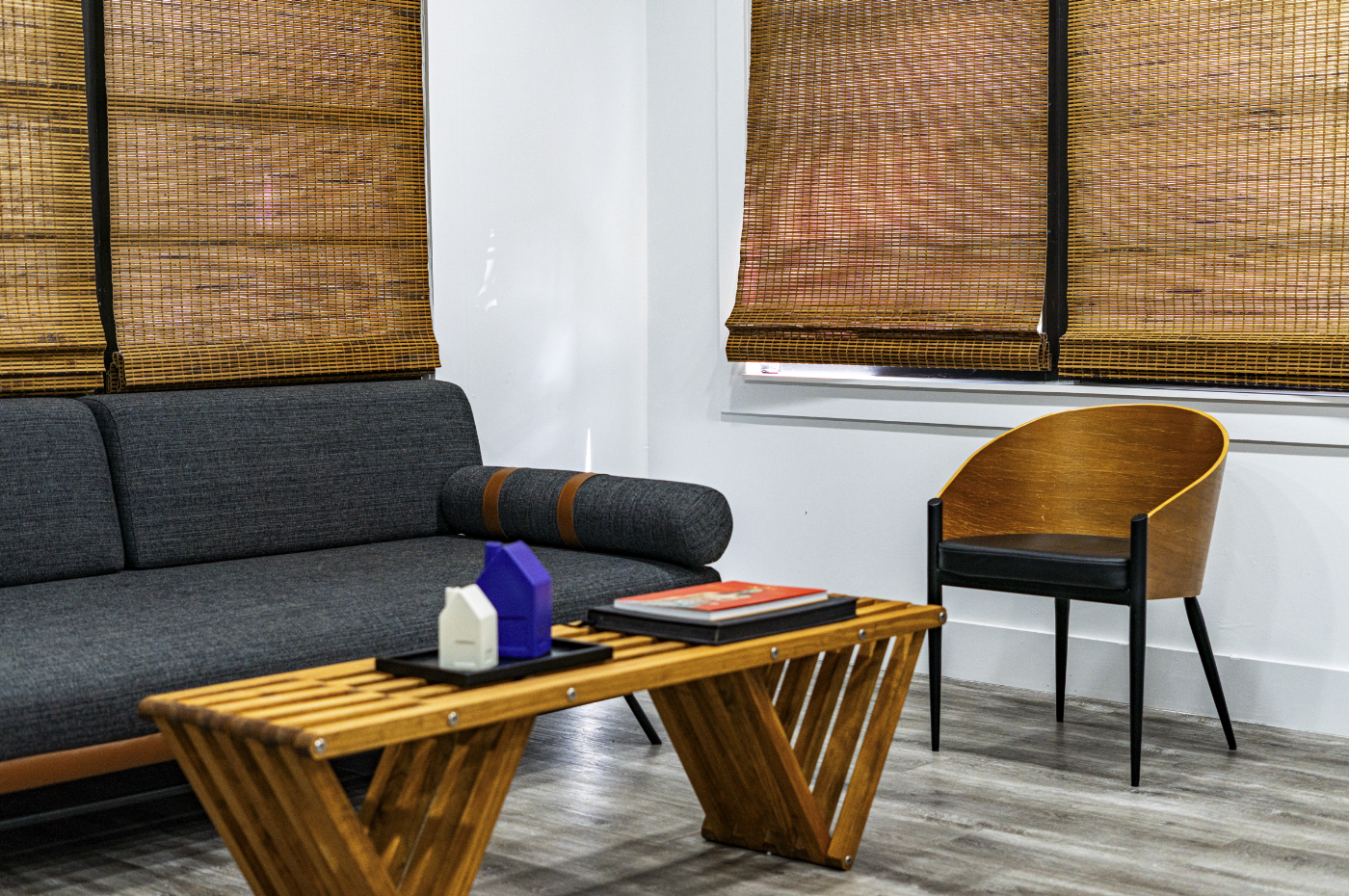 A modern living room with a dark gray sofa, a wooden slatted coffee table, a wooden accent chair, and bamboo window shades.