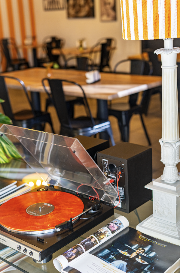 A close-up of a vintage turntable playing a record with a transparent dust cover, with a speaker beside it, in a room with a wooden dining table, black chairs, and wall art in the background.