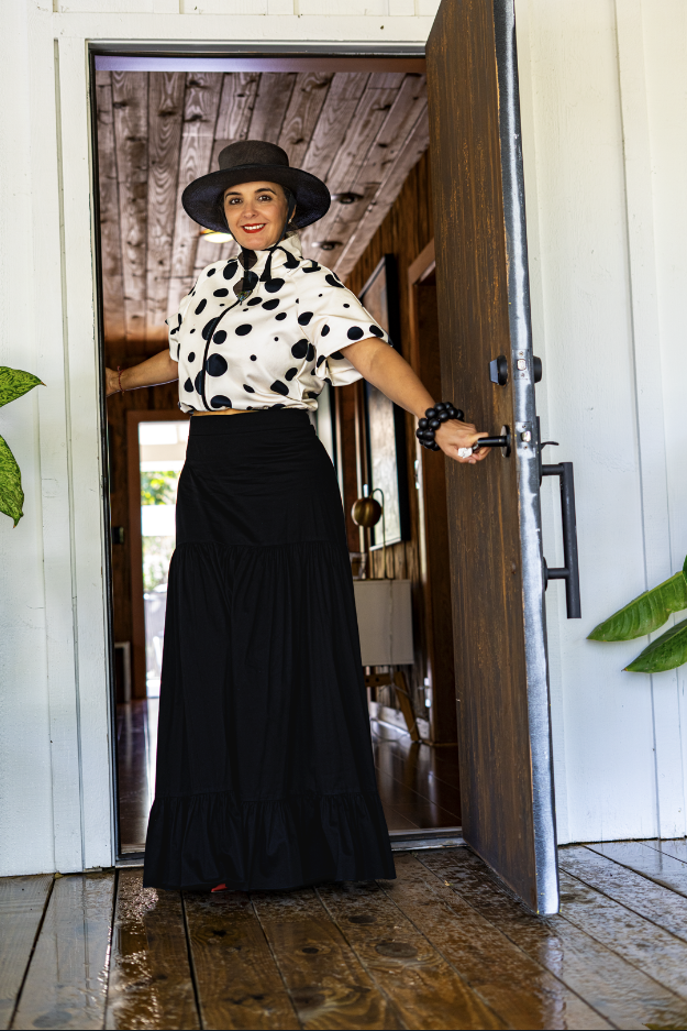 A woman standing in a doorway, smiling, wearing a large black hat, a white blouse with black polka dots, a long black skirt, and a black bracelet.