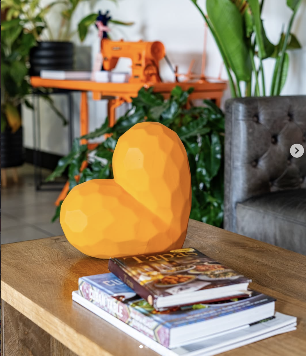 A yellow, faceted heart-shaped lamp on a wooden table with magazines underneath. In the background, there are green plants, a gray upholstered chair, and a workspace with an orange sewing machine.