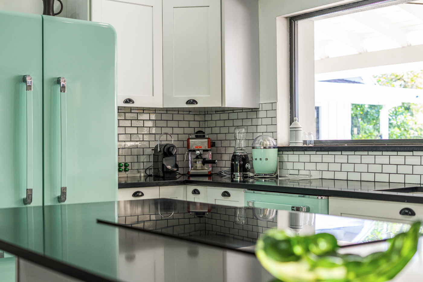 A kitchen with white cabinets, a black countertop, and a black and white subway tile backsplash. On the counter, there are small kitchen appliances including a coffee machine, espresso machine, blender, and a vintage turquoise cooler. There's a windo