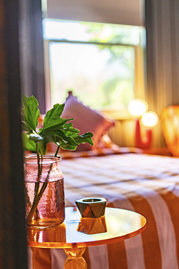 A bedroom with a window, a bed with striped bedding, a pink glass vase with green leaves on a small orange side table, and blurred warm lights in the background.