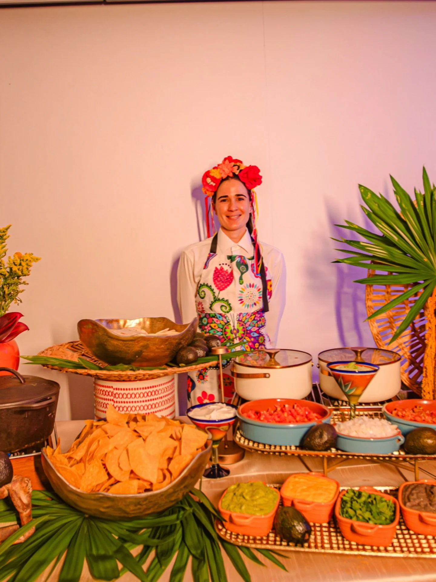 A woman dressed in traditional Mexican attire with a colorful floral headpiece, standing behind a table of Mexican food and decorations.