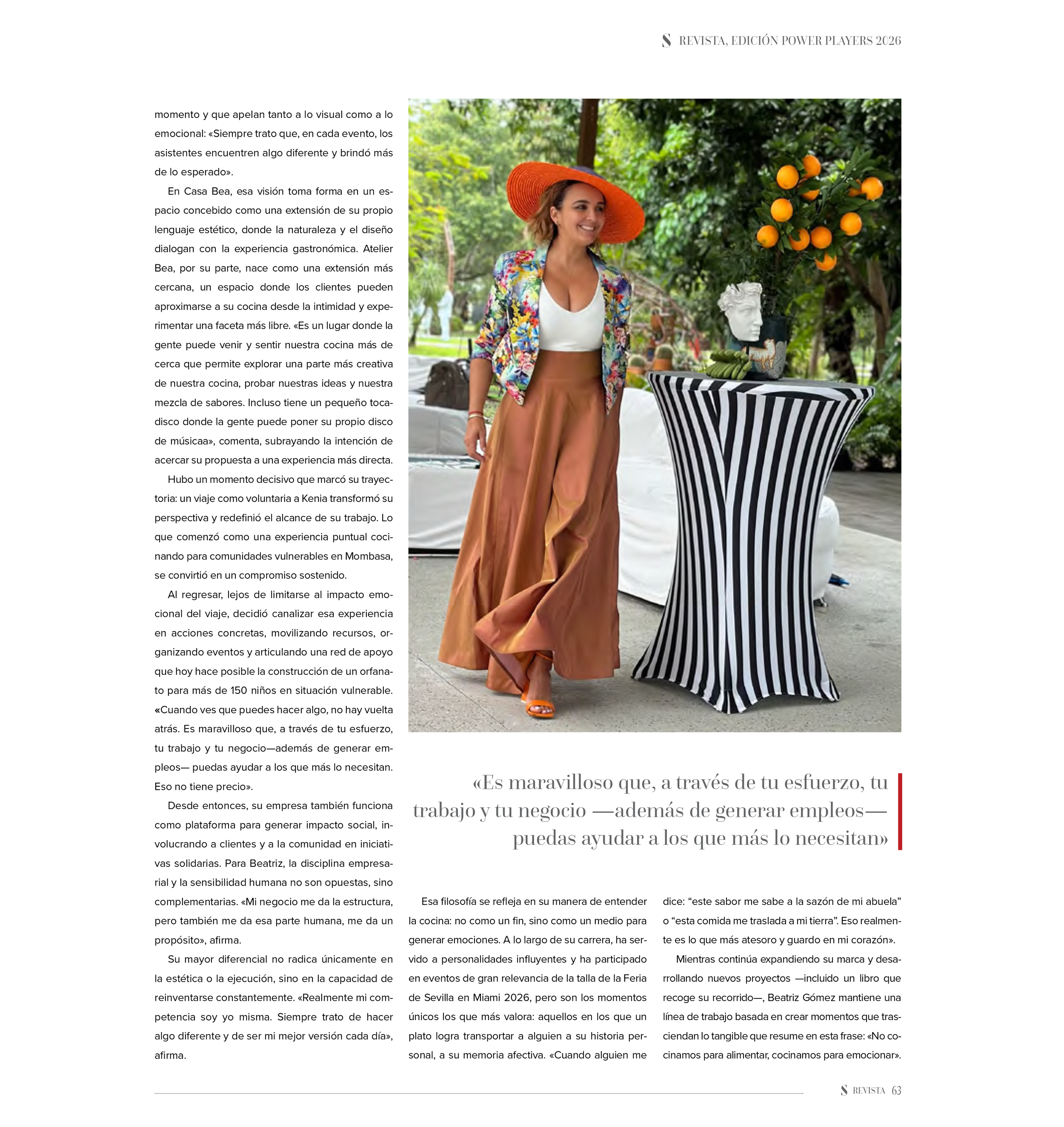 Woman in colorful blazer, white top, and brown wide-leg pants with orange hat standing next to a black and white striped cocktail table with orange fruits and a white sculpture on top, outdoors with greenery.