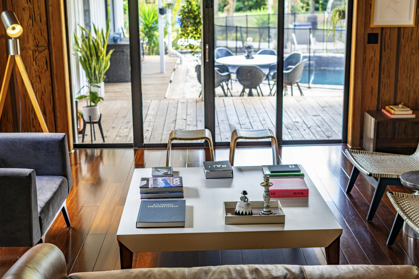 Living room with a white coffee table, books, and decorative items, adjacent to large glass sliding doors leading to an outdoor wooden deck with a round table and chairs.