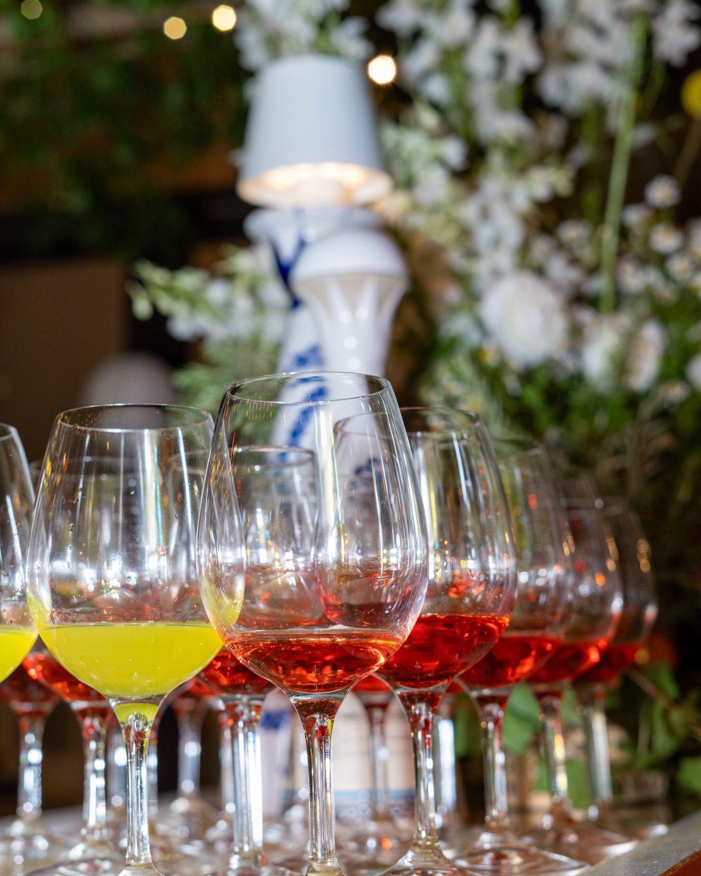 Multiple wine glasses filled with yellow and red wine arranged on a table with a blurred background of flowers and a white lamp.