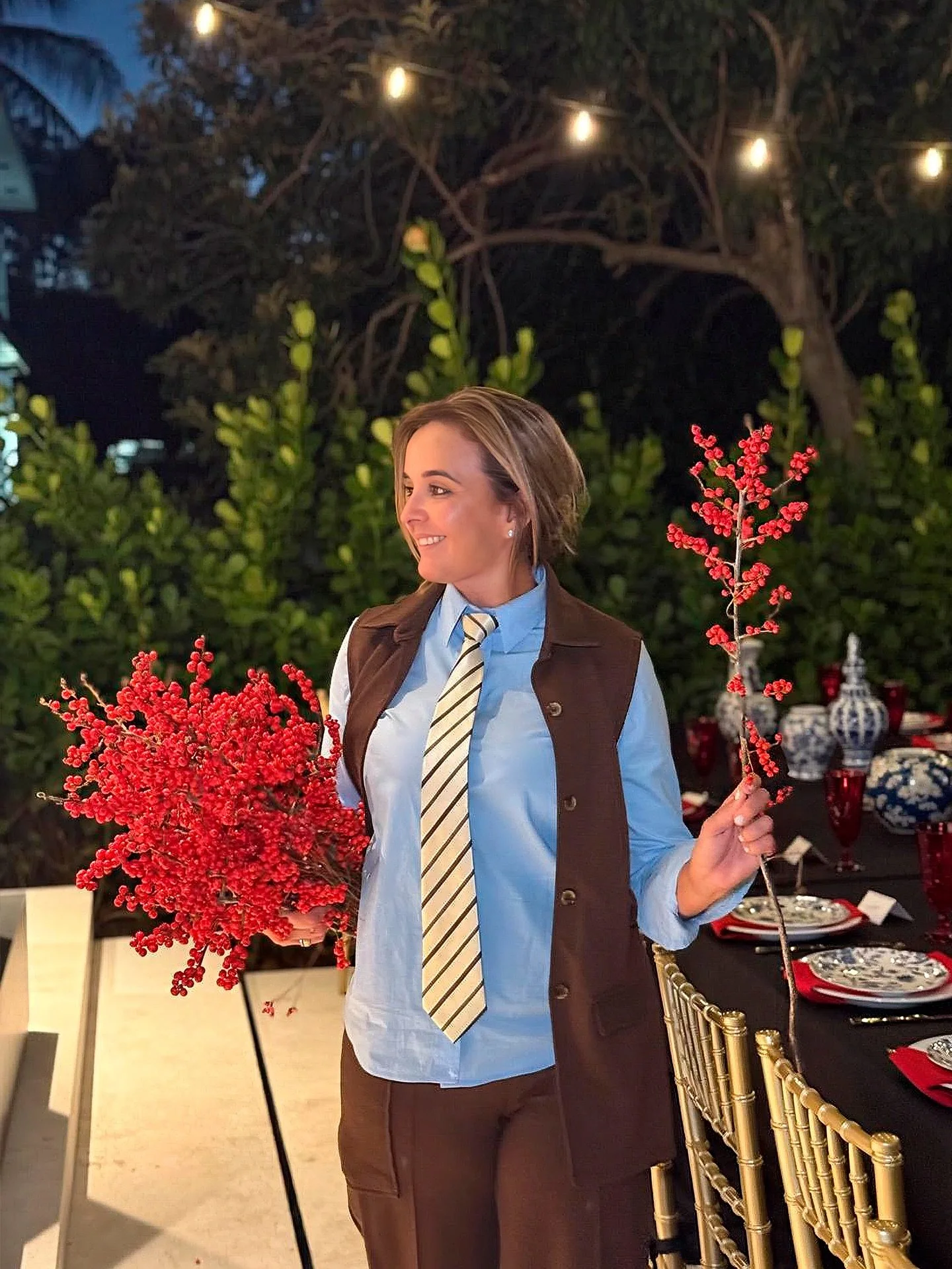 Woman holding red berry branches at an outdoor event with elegant table settings and fairy lights in the background.