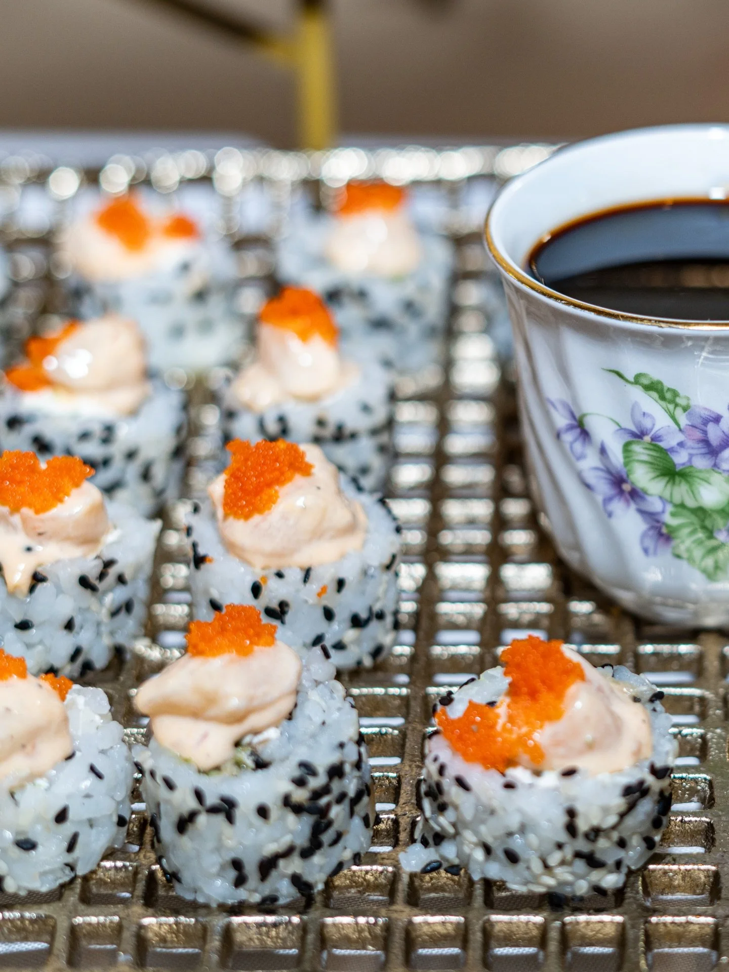 Sushi rolls topped with orange fish roe and a dollop of creamy sauce, with a cup of black coffee in a floral mug in the background.