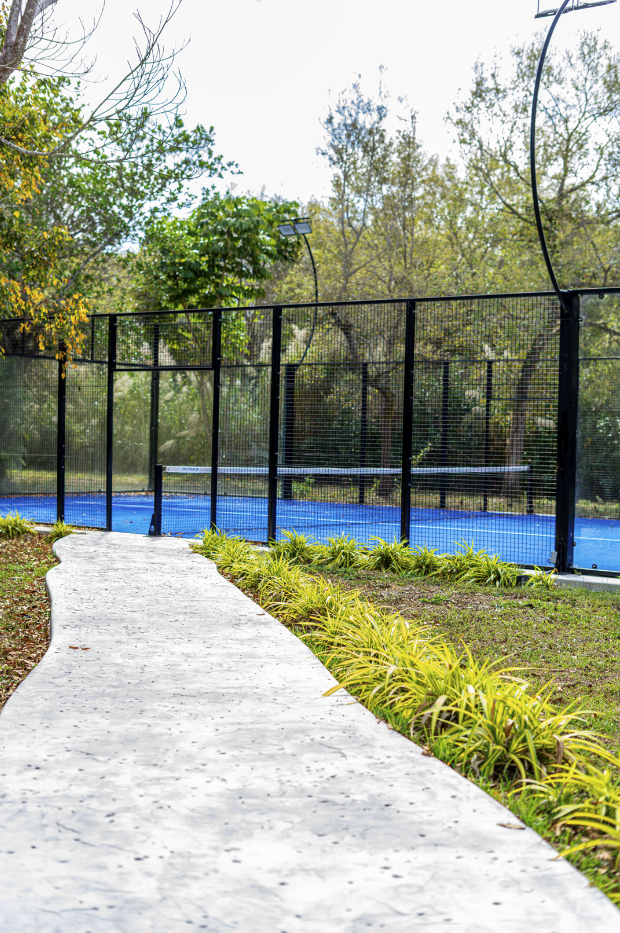 Concrete pathway running alongside a tennis court with black fencing, surrounded by green trees and bushes.