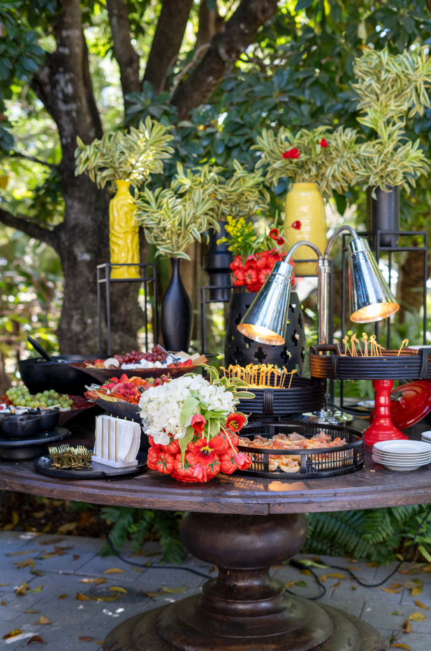 Outdoor buffet table with food, flowers, and decorative vases, set against trees and greenery.