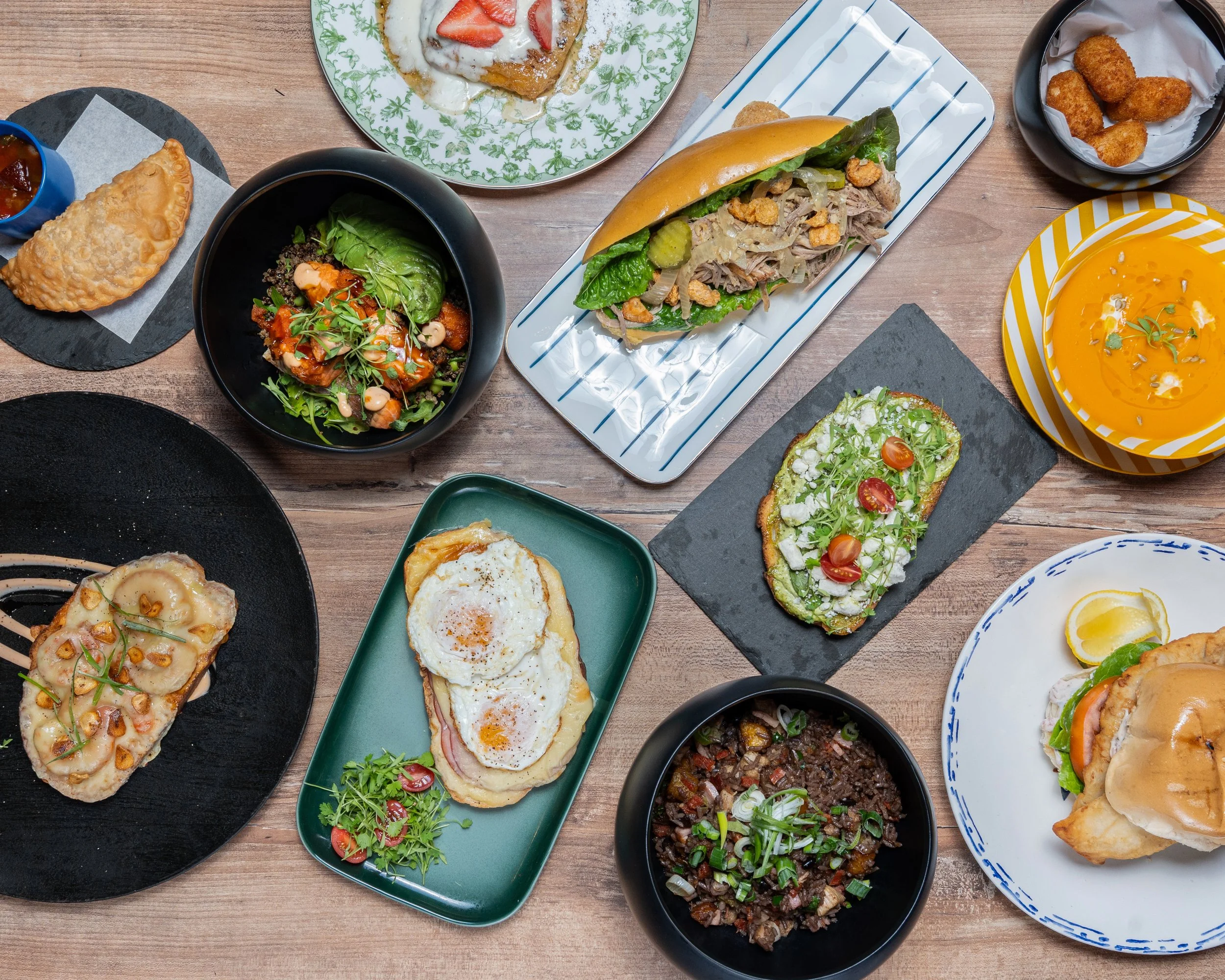 Assorted dishes including a sandwich, salad, pizza, fried food, soup, tacos, and other breakfast items on various plates and bowls on a wooden table.