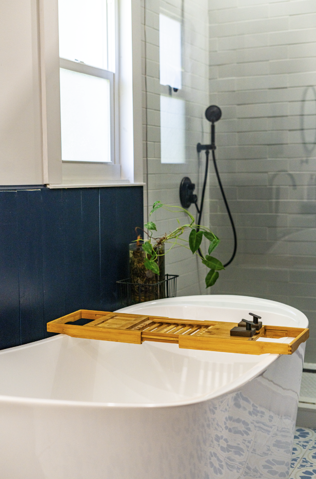 A bathroom with a white bathtub, a bamboo bath tray, a potted plant, and a walk-in shower with black fixtures and a window.