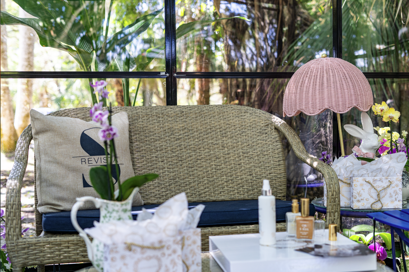 A wicker outdoor sofa with a beige cushion and a decorative pillow on it, surrounded by flowers, a pink scalloped lampshade, and skincare items on a white table, with a glass window and lush greenery outside.