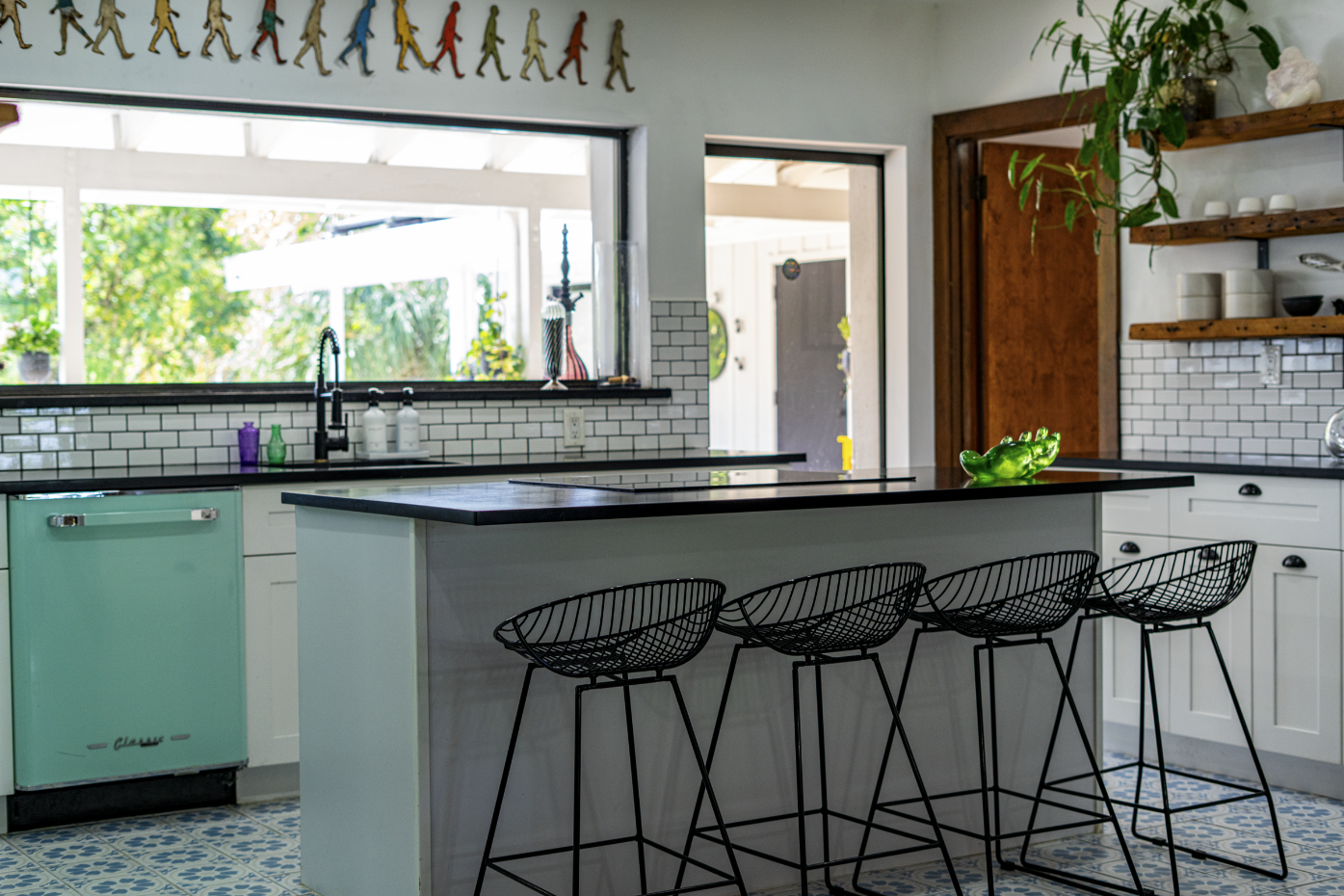 Modern kitchen with white cabinets, black countertops, and wire bar stools at a kitchen island. Decor includes plants, vases, and a colorful art piece along the ceiling. Large window and door leading outside, with a tiled floor.