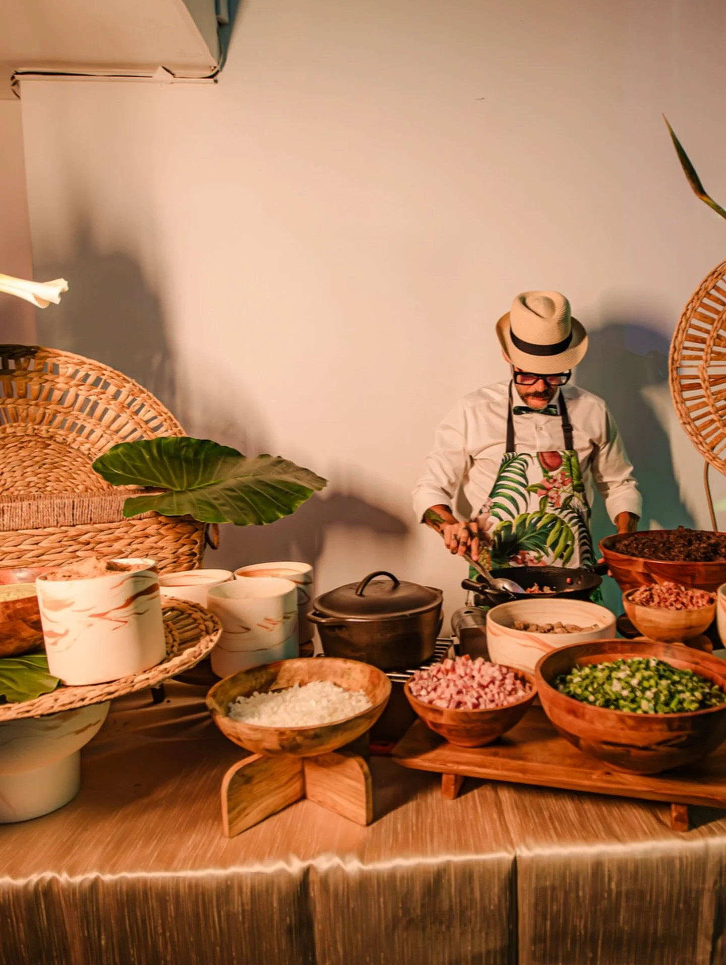 A man wearing a hat, glasses, and an apron is cooking at a food station decorated with tropical leaves and wicker baskets, surrounded by bowls of chopped vegetables and rice.
