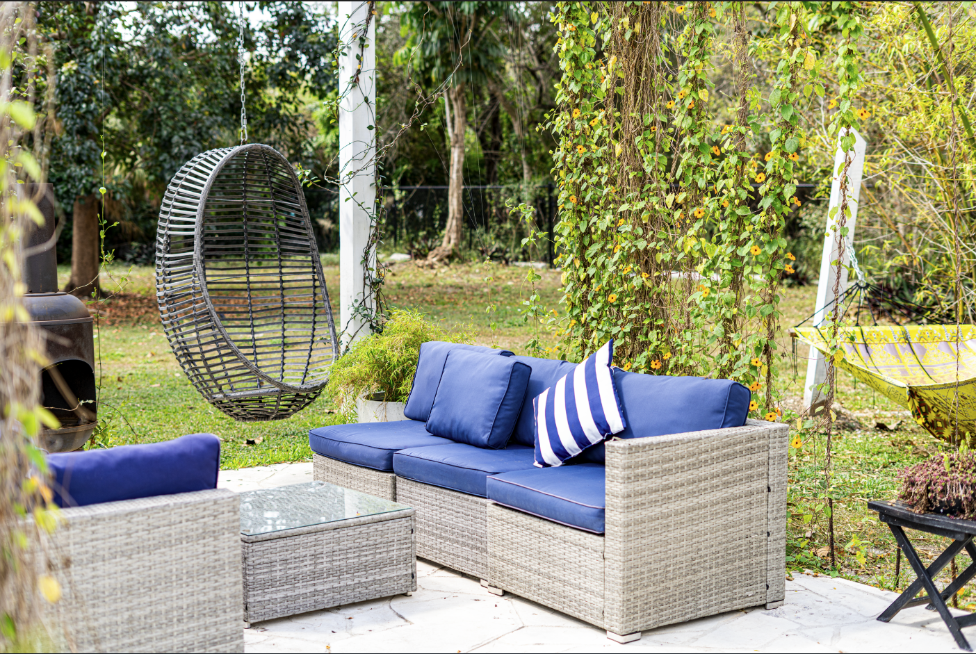 Outdoor patio with blue cushioned wicker furniture, a glass-top coffee table, a hanging egg-shaped chair, and greenery including trees and climbing plants with yellow flowers.