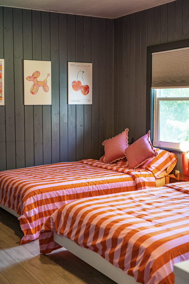 Two beds with orange and white striped bedding and pink pillows in a room with dark wood-paneled walls, posters of cherries and a balloon animal, and a window with a view of greenery outside.