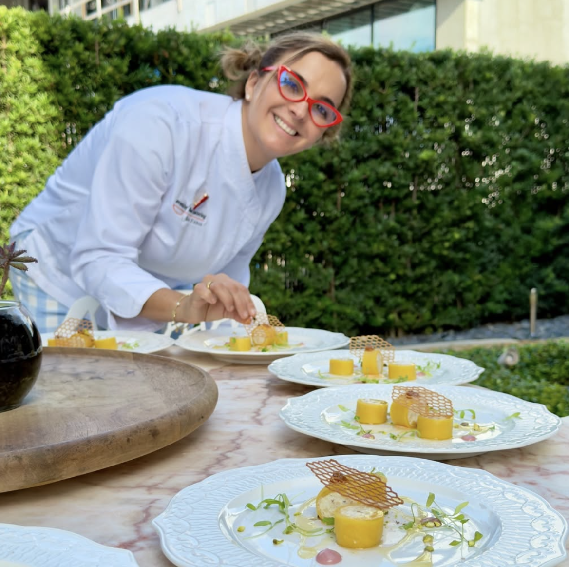 A woman with red glasses and a white chef's coat is smiling and garnishing plates of yellow food, possibly desserts, on an outdoor table with greenery in the background.
