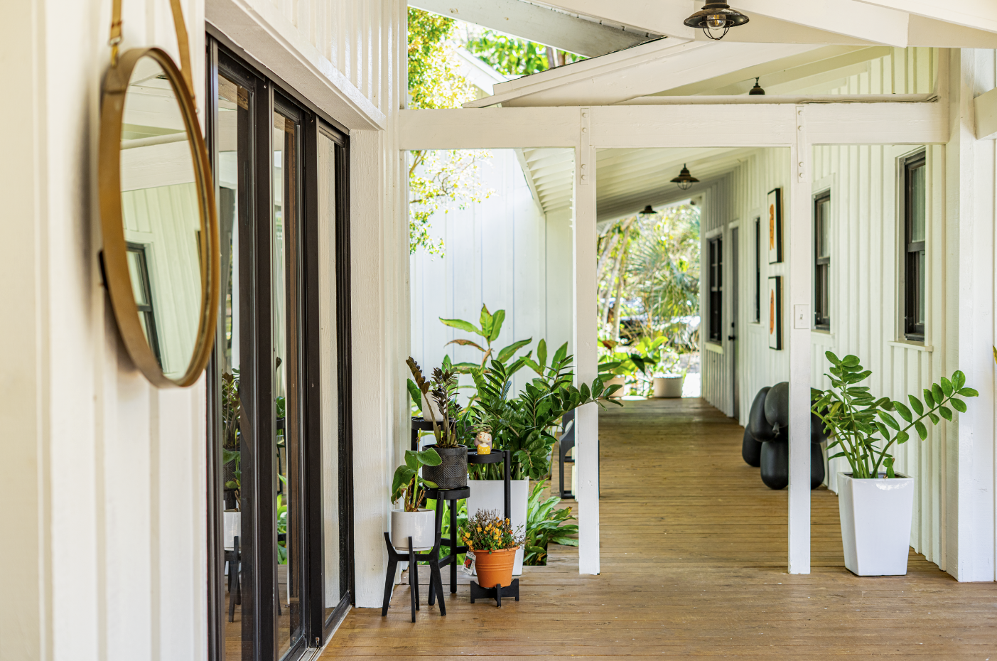 Covered porch area with potted plants, black chairs, and framed pictures on the wall, overlooking a garden with trees and greenery.